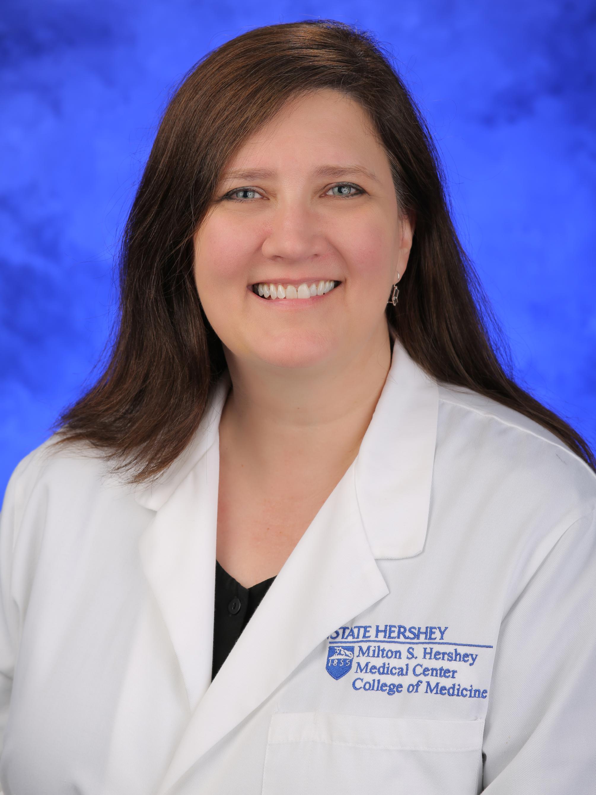 Dr. Shelly D. Timmons poses in a white physician's coat, against a blue, lightly textured backdrop.