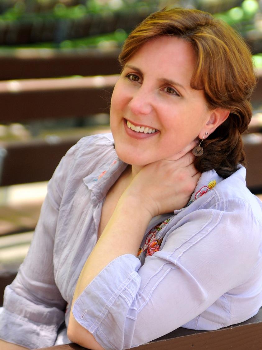 A woman smiles and rests her left hand on her neck.