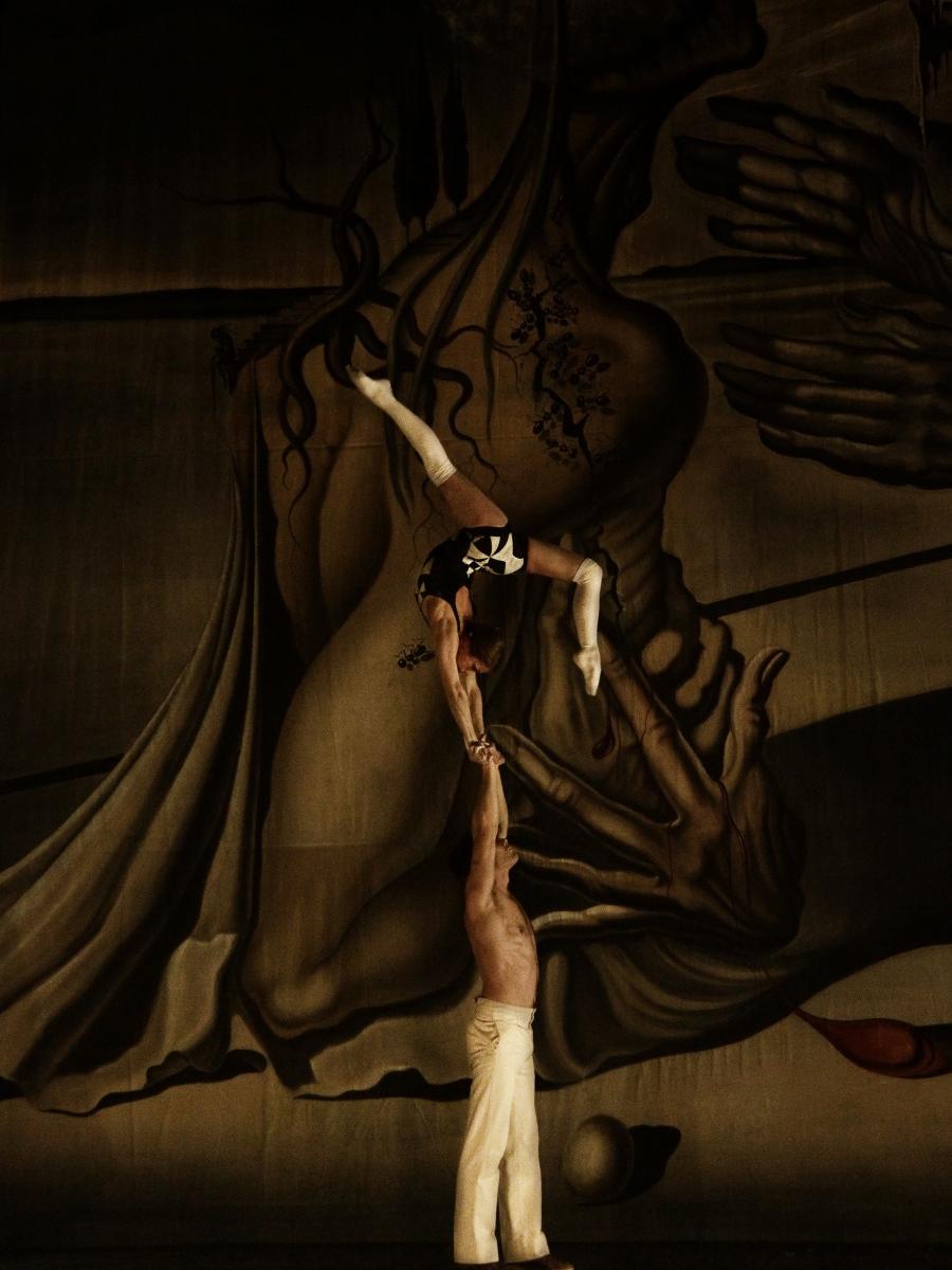 A male acrobat extends his arms straight above his head to hold up a woman, who bends her right leg back behind her and extends her left straight up in a scene from Compagnia Finzi Pasca’s production of “La Verità.”