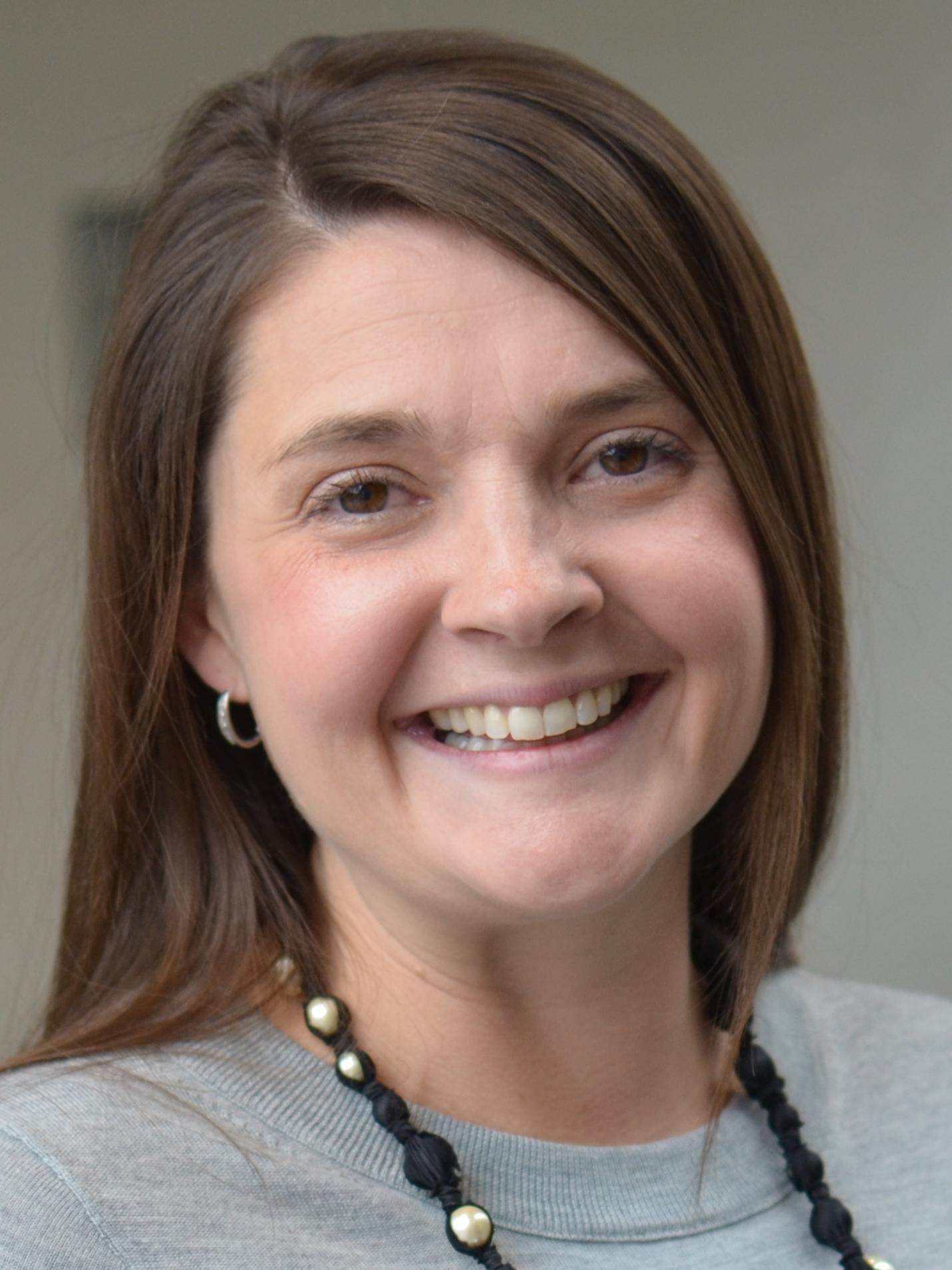 A woman with long brown hair smiles in front of a gray background. Her shirt is gray and she is wearing silver hoop earring in her right ear and a black beaded necklace with white beads throughout 
