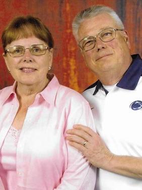 A woman in a pink shirt and glasses and a man in a white Penn State polo shirt with blue accent colors and glasses smile at the camera.