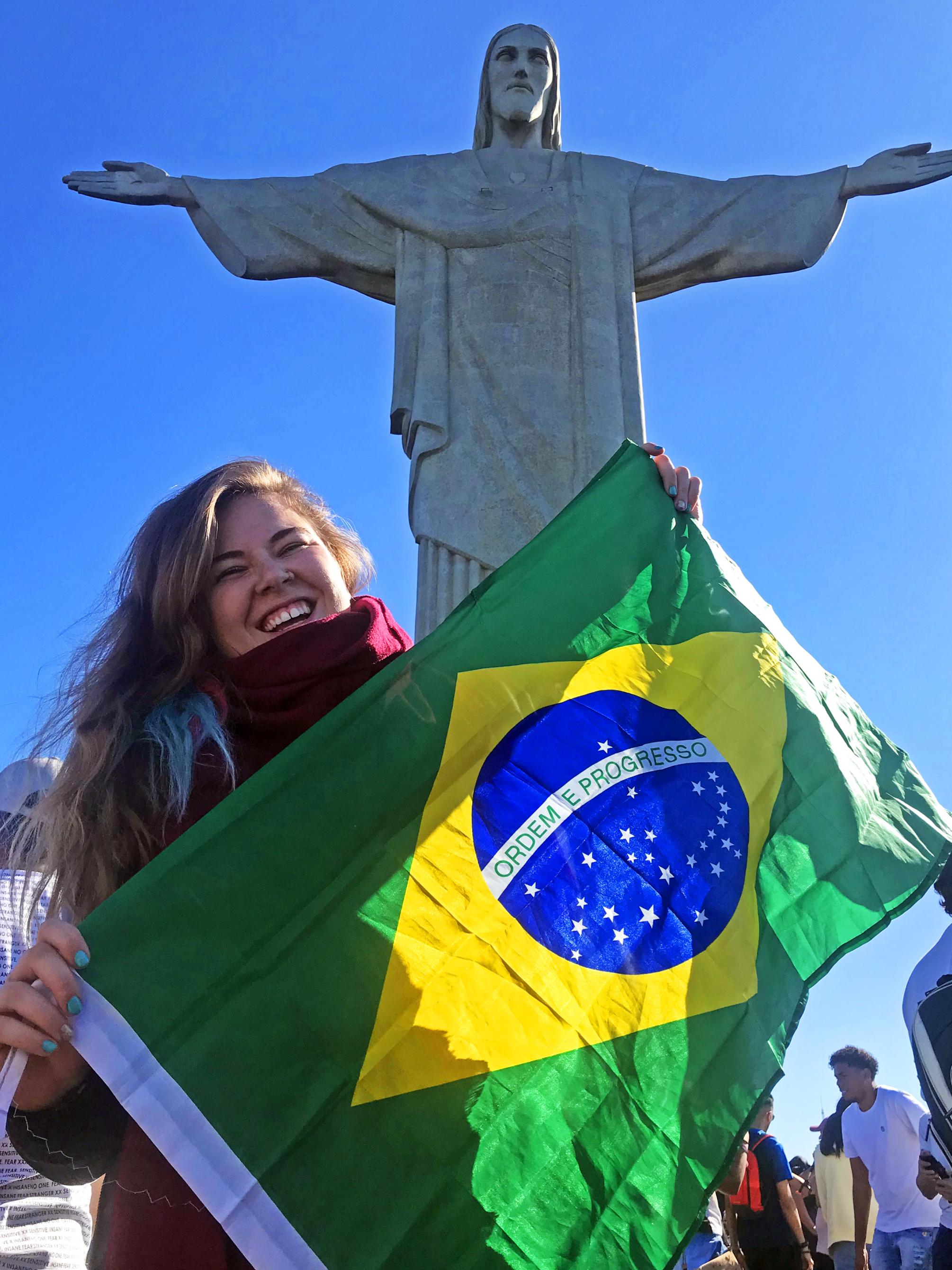 A young woman stands by the Christ the Redeemer statue in Brazil, holding a Brazilian flag