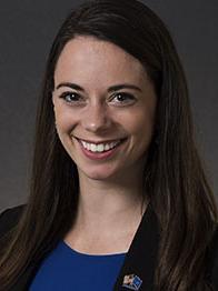 Headshot of a young woman with brown hair wearing a blue shirt and black jacket 