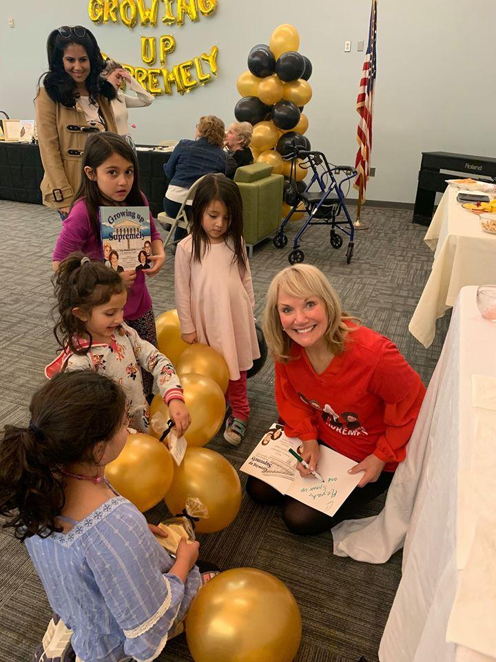 woman sitting on ground signing a book with young kids surrounding her