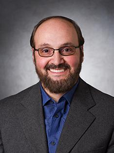 A headshot of a man in a blue button-down shirt, a suit jacket and glasses smiling at the camera in front of a gray background