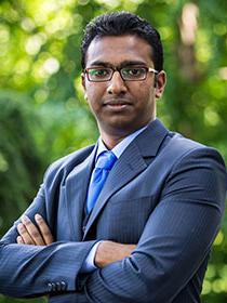 A man in a suit, blue tie and glasses poses with his arms crossed in front of a blurred green background. 