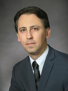 A headshot of a man wearing a gray suit jacket, white shirt and a dark gray tie 