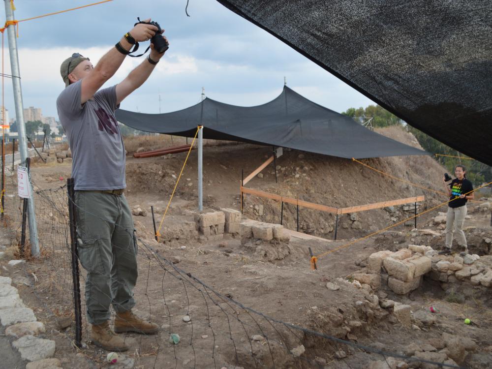 Tim Hay raising his arms to take a photograph of Tel Akko dig site