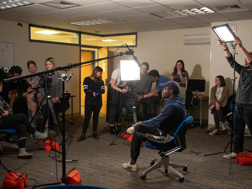 Students on a video team practice setting up lights and camera equipment around a single subject