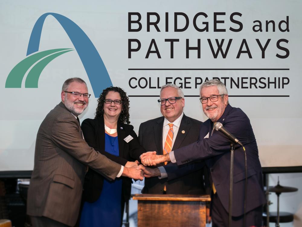 Four college presidents shake hands on stage.