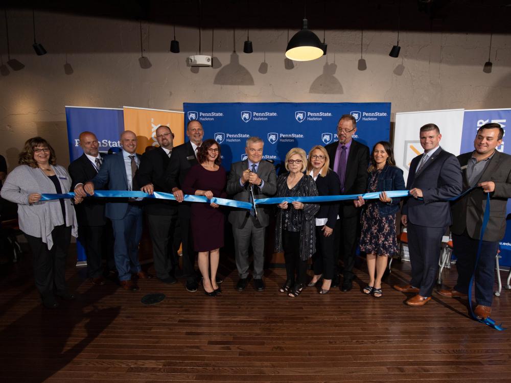 A group of eleven people lined up and holding a ribbon with one person cutting it. 