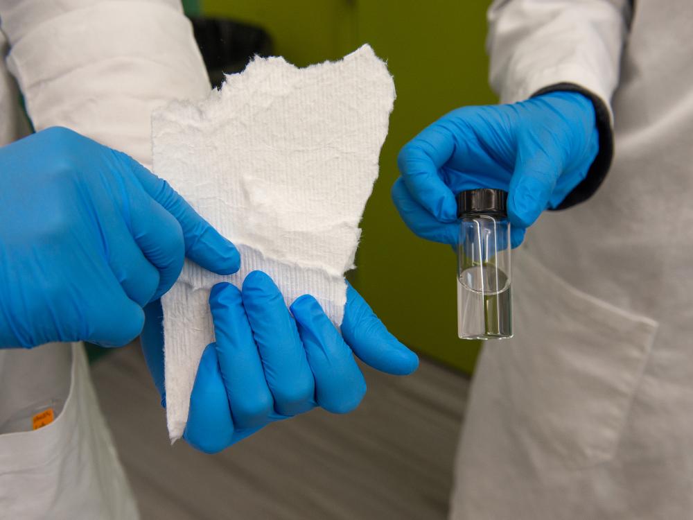 Hands in blue lab gloves hold a paper towel and a small vial of liquid. 