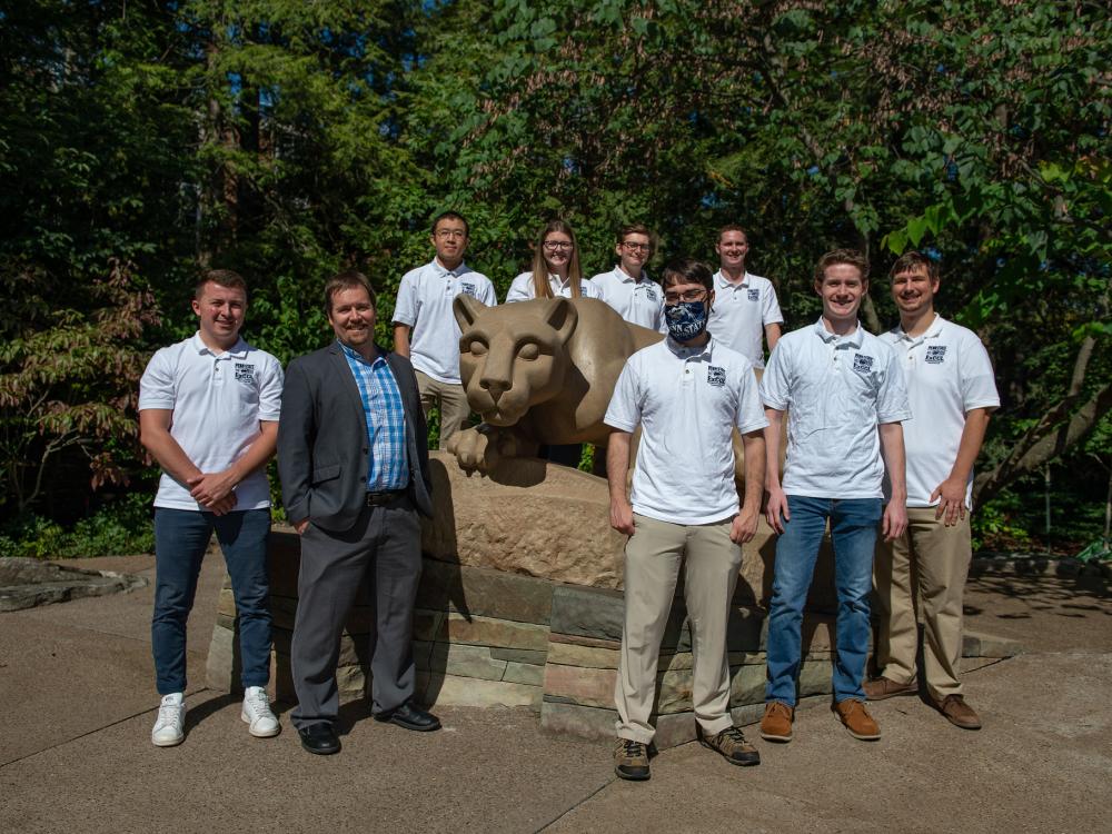 A professor and a group of students wearing white polo shirts pose in front of the Nittany Lion shrine. 