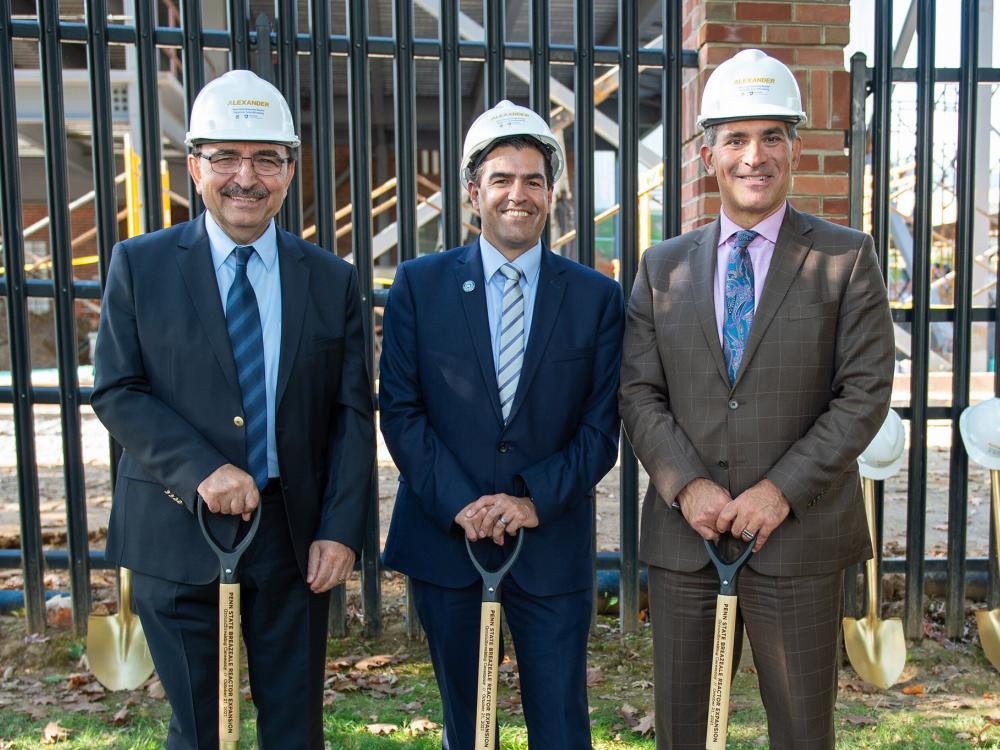 Three men wearing suits and hard hats pose with shovels in front of a blank fence.