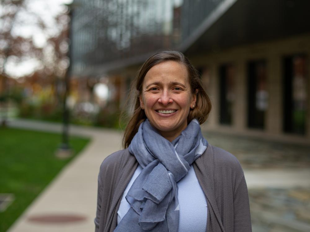 A researcher wearing a gray cardigan and blue scarf smiles with an engineering building and sidewalk in the background