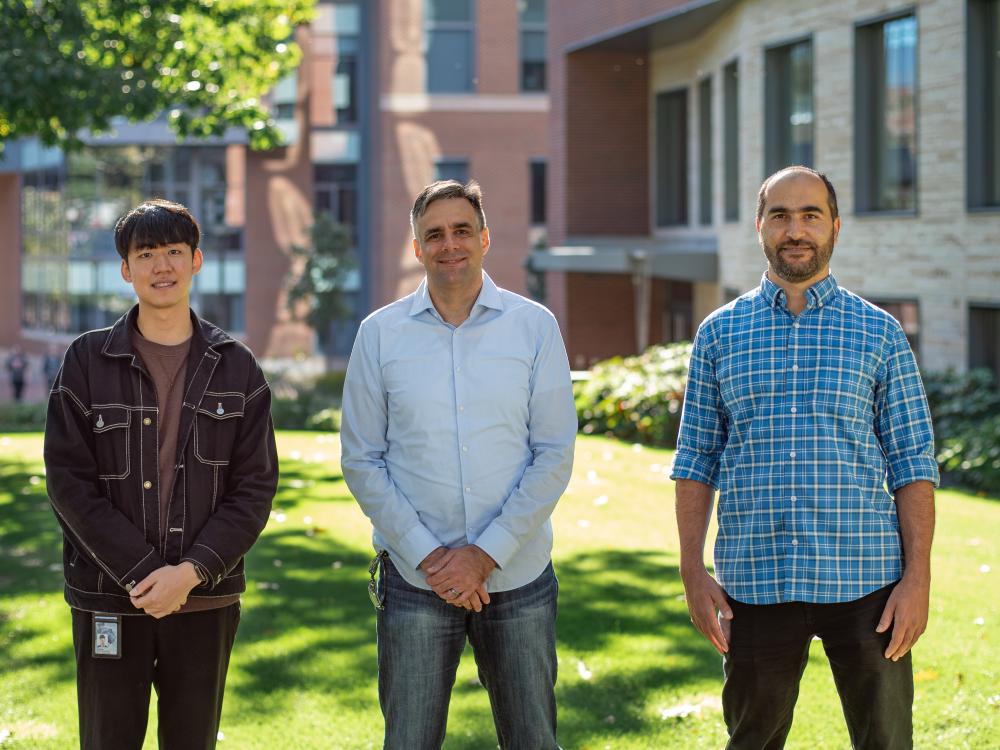 Three men stand outside on a sunny day with a brick building in the background.