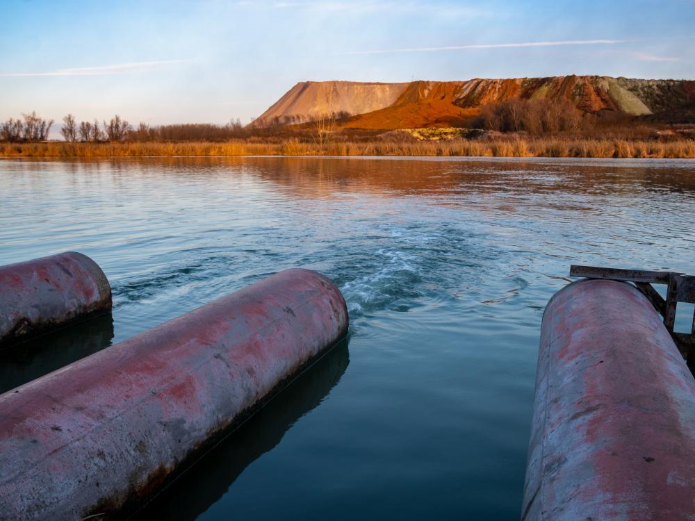 Three rusted pipes outputting into a lake with red and white hills in the distance.