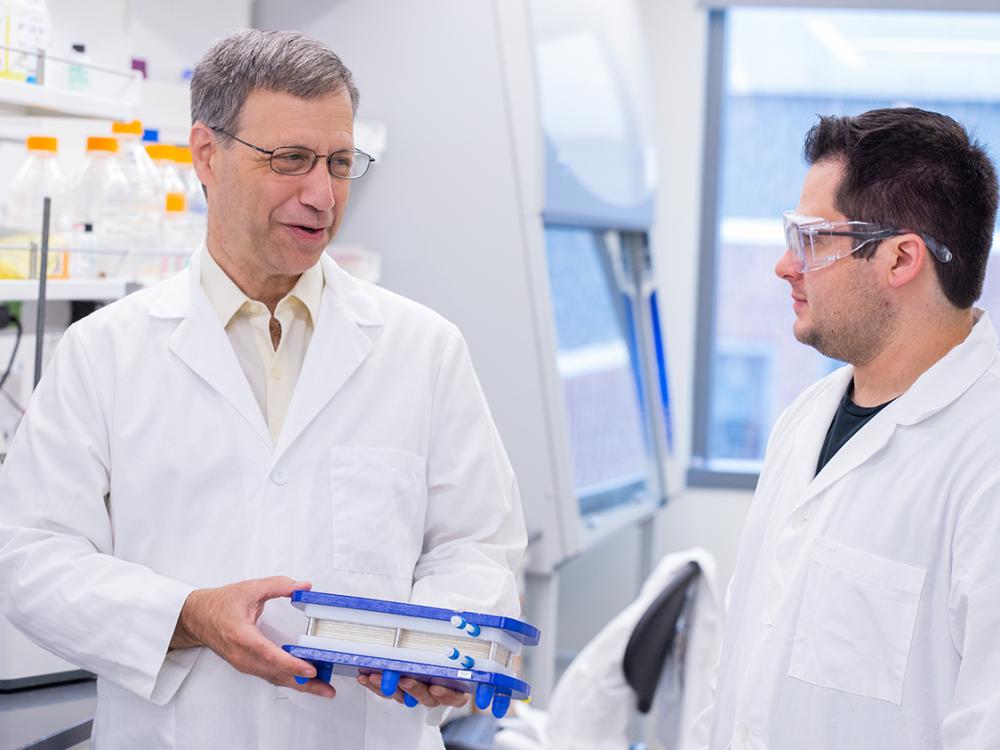 A man smiles and holds a membrane model while a male student in a lab coat looks on.