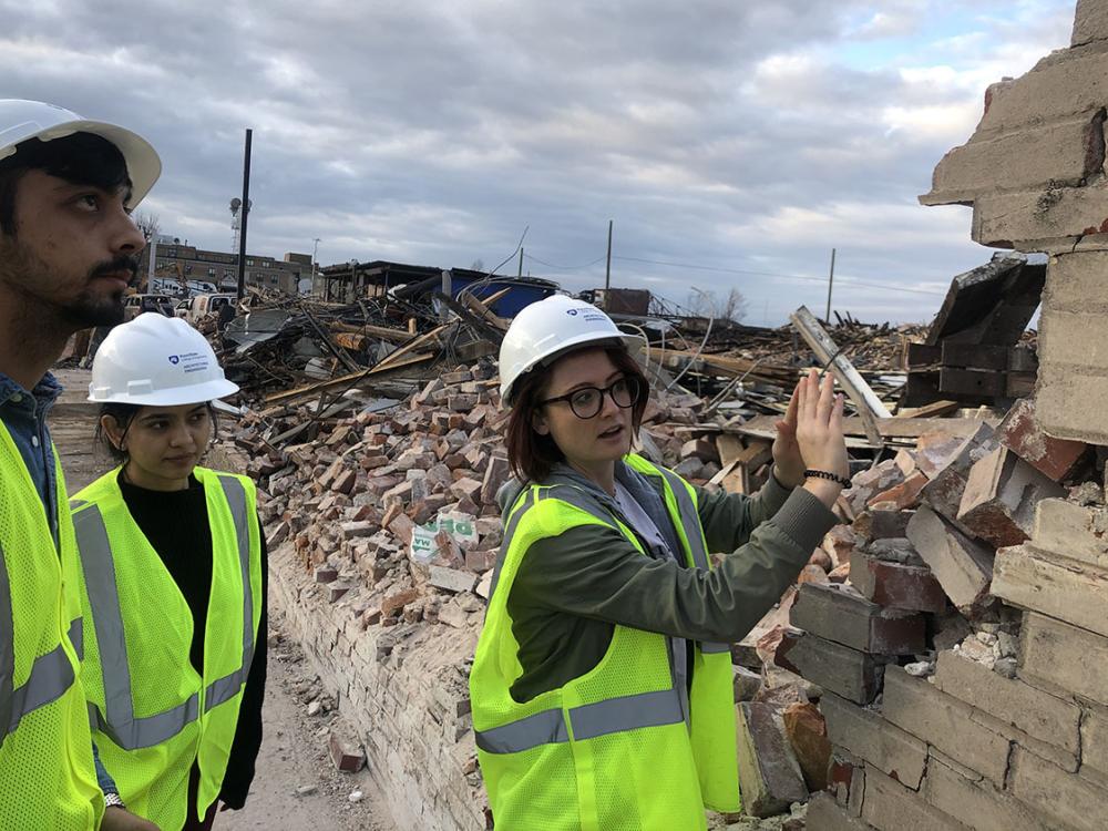 Three people wearing hard hats and neon vests stand near a mostly demolished brick wall as one of the people gestures at the wall in explanation to the other two people who look on. 