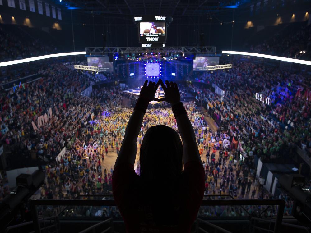 In the foreground a person is silhouetted as they hold up their hands in a 'diamond' formation. THON 2019 at the Bryce Jordan Center is in the background.