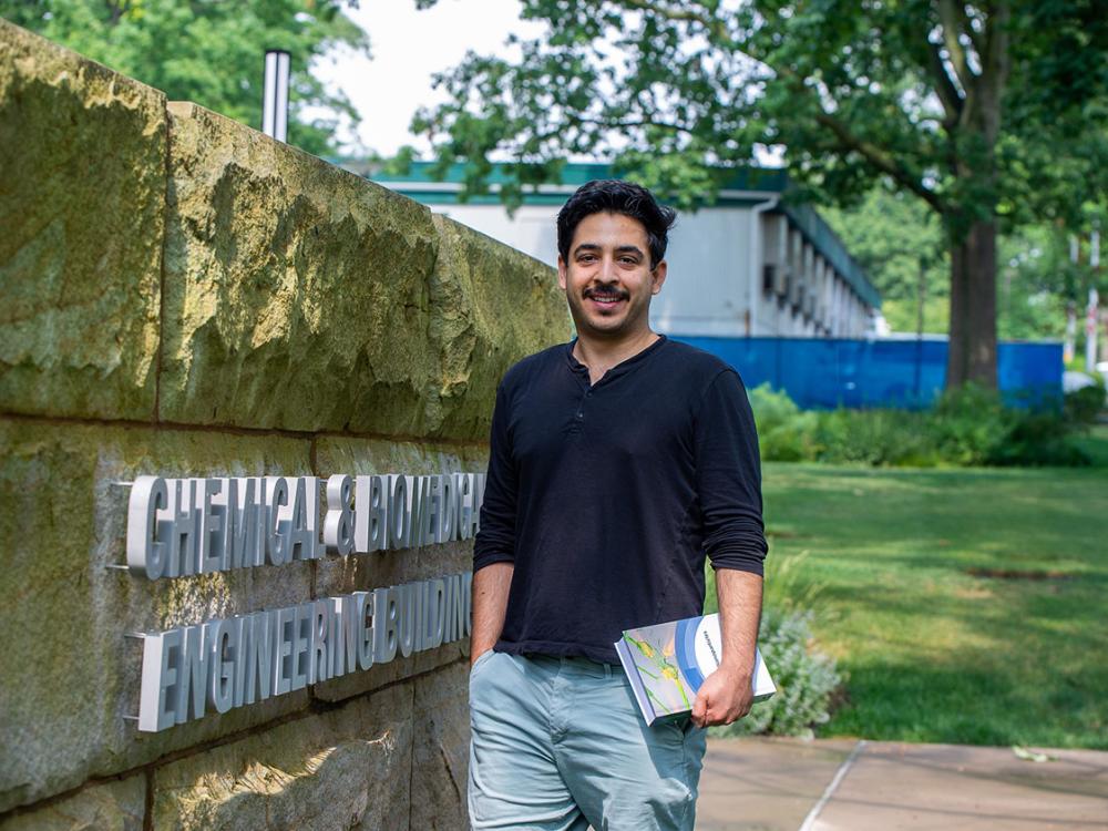 A person wearing a dark long sleeved shirt holds a textbook in front of a "Chemical and Biomedical Engineering Building" sign on a stone wall outside