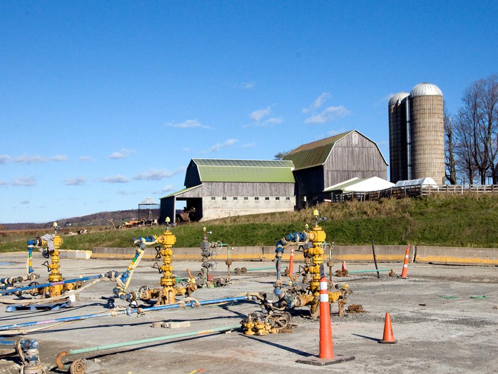 A gas well with a barn and corn silos in the background