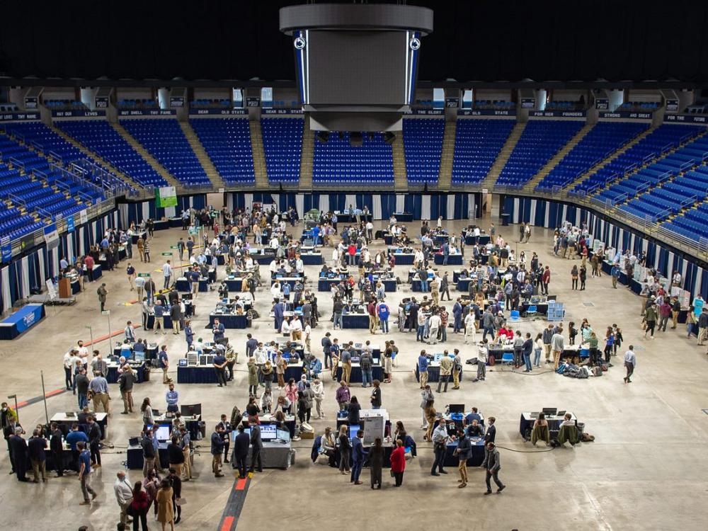 Photo taken from stands shows nearly eighty display tables set up in a large arena surrounded by people presenting and talking.