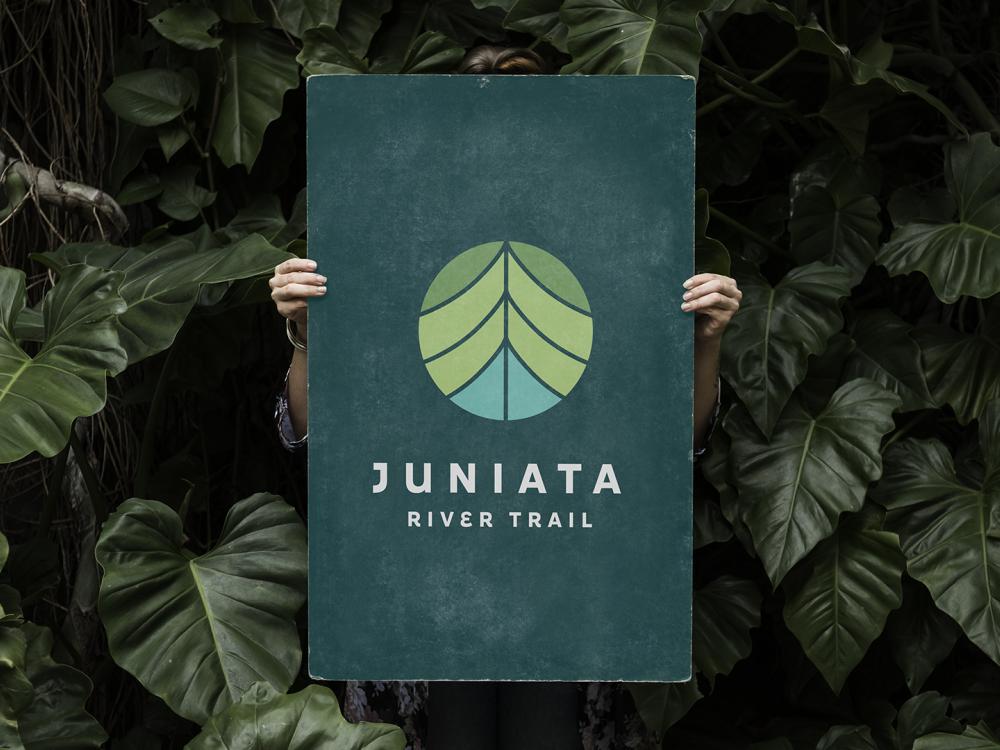 Someone holding up a sign for the Juniata River Trail in front of their body among a background of plants.