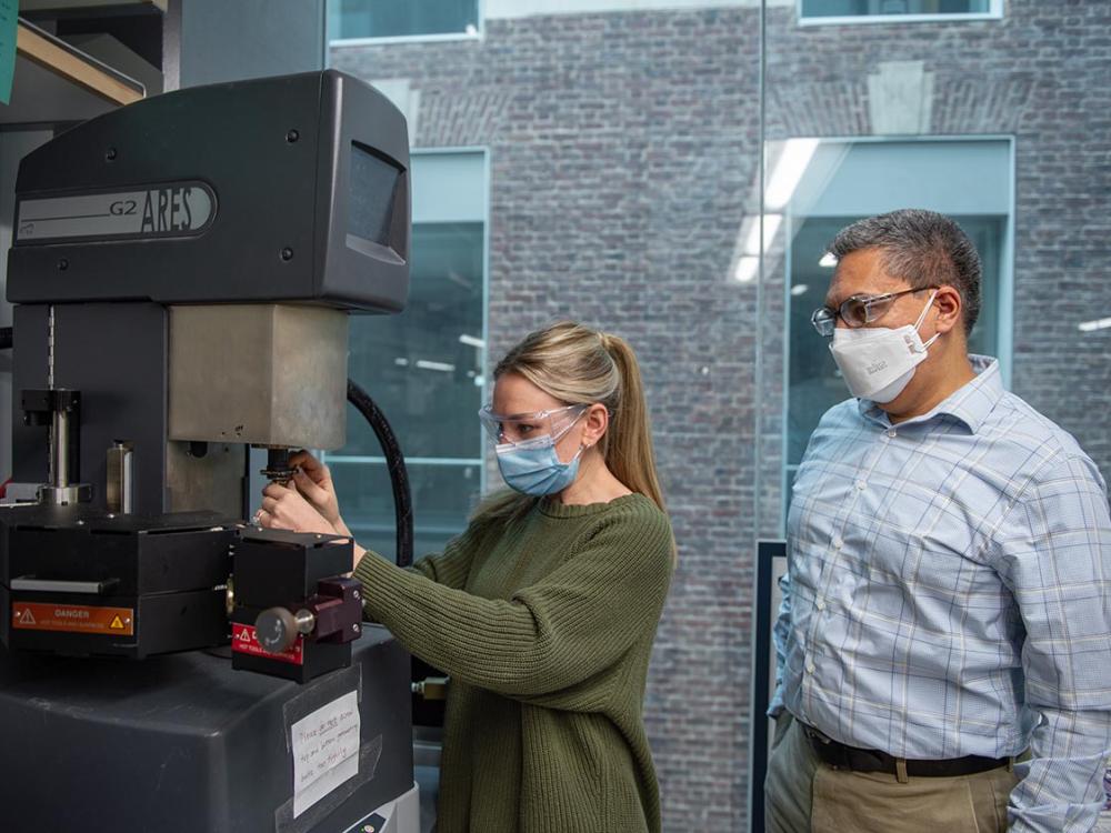 A woman wearing lab glasses adjusts a machine while a man stands near her looking on.