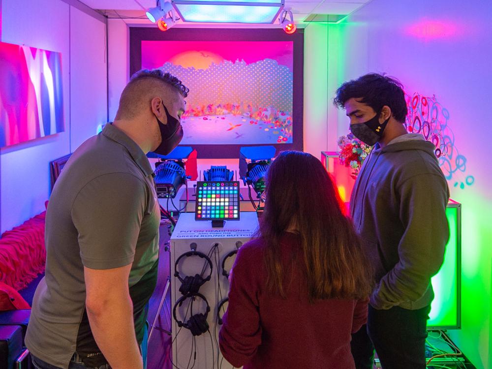 Three people study a display screen in a technical lab with brightly colored lights.