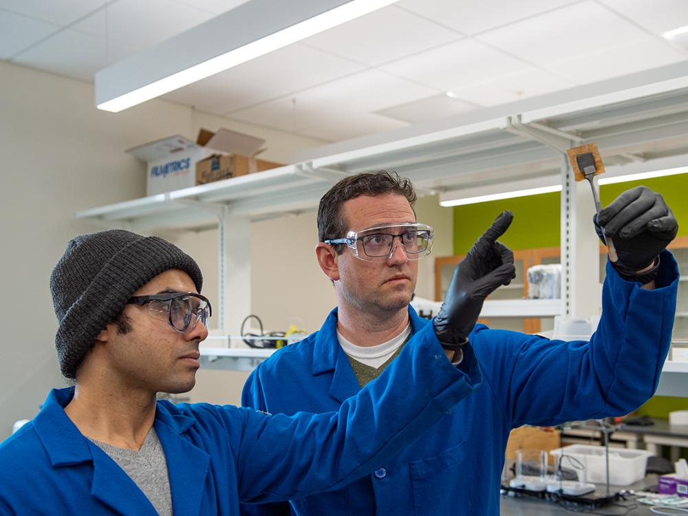 Two men in lab coats and protective glasses look up at a yellow, square membrane sample held by a lab tool resembling tweezers.