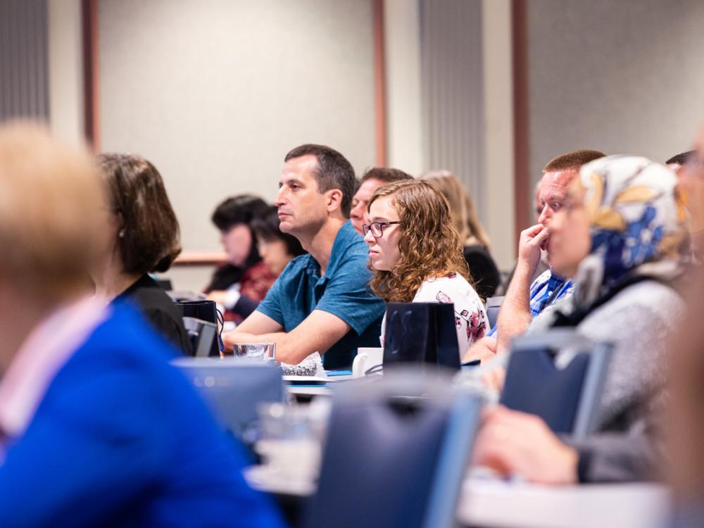 Many people sit in a lecture hall with laptops looking toward the front of the room