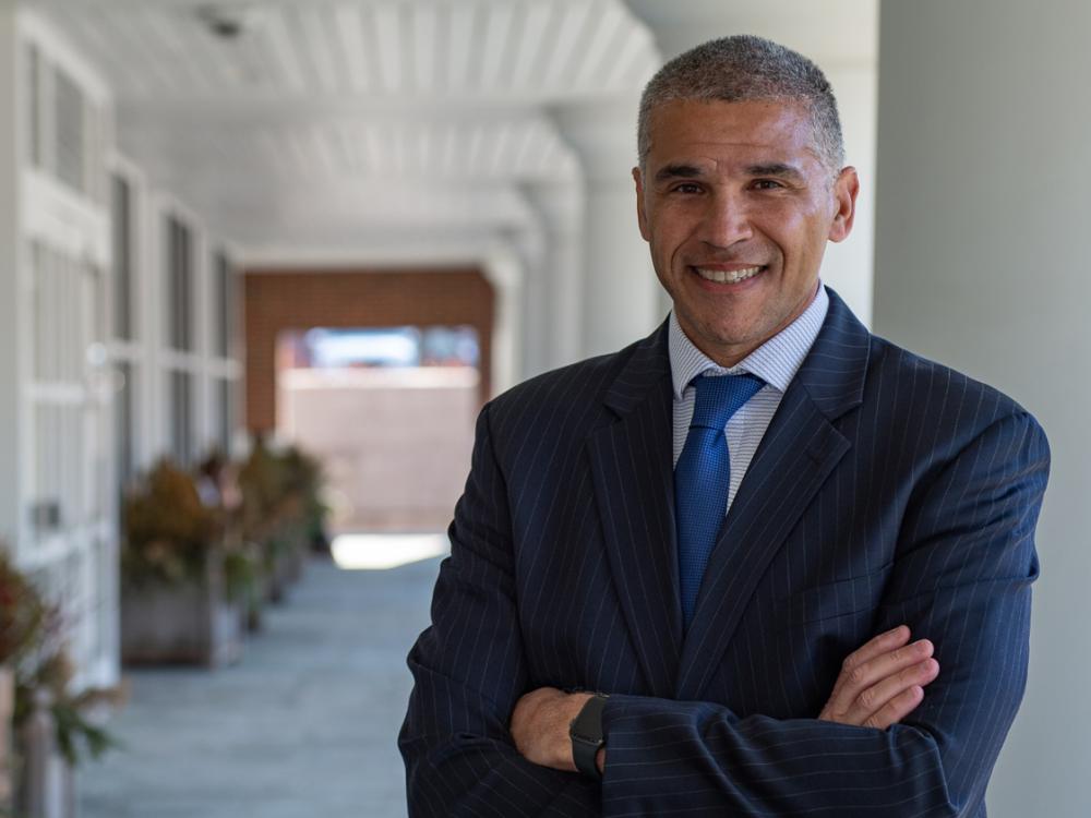 A smiling man in a suit and with his arms crossed stands in a covered walkway. 