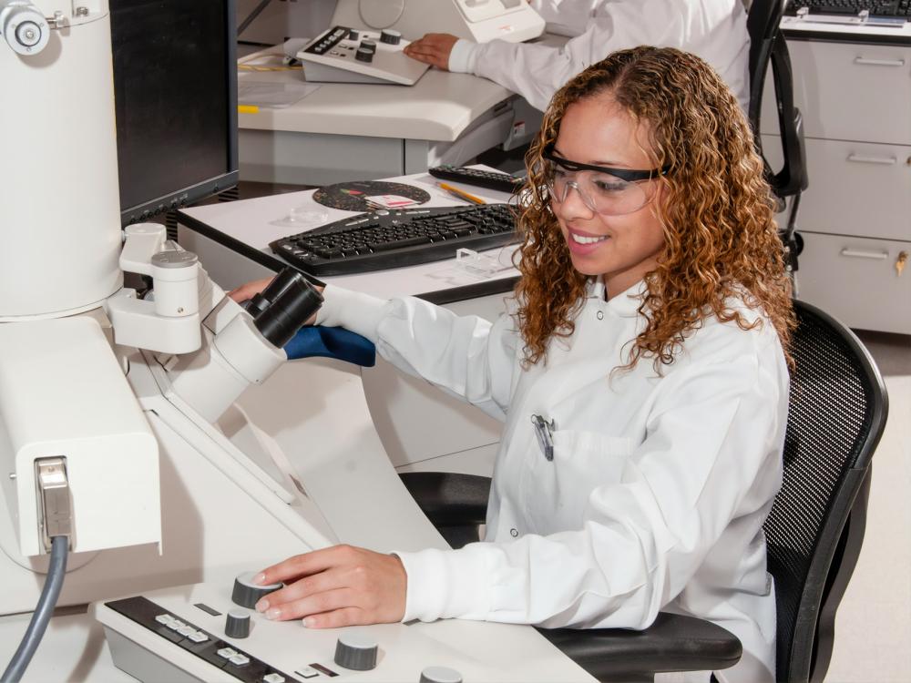 A person in a white lab coat and safety glasses works at a lab station.