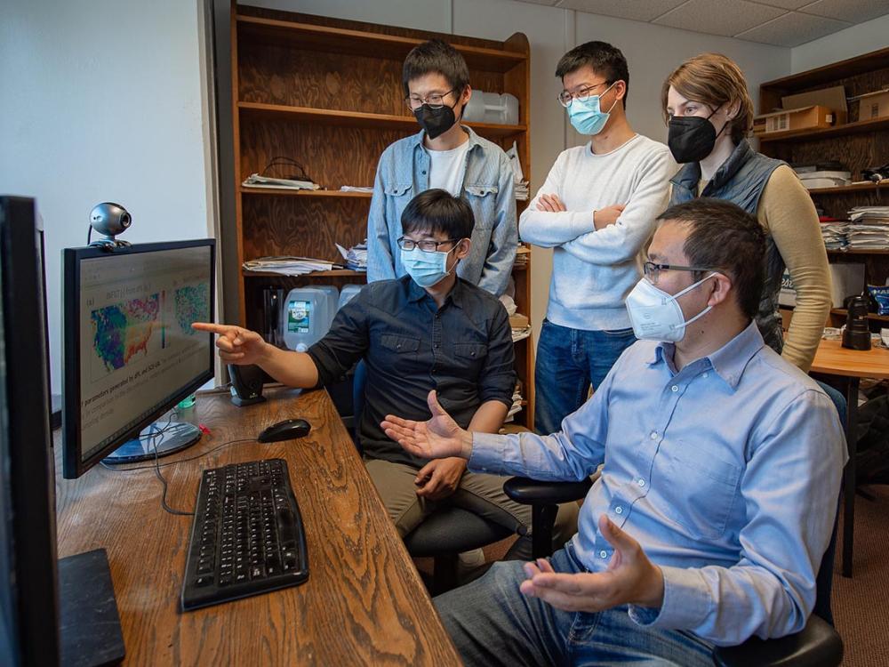 Five researchers wearing masks stand and sit around a computer, where two maps of the United States are displayed on the monitor. 