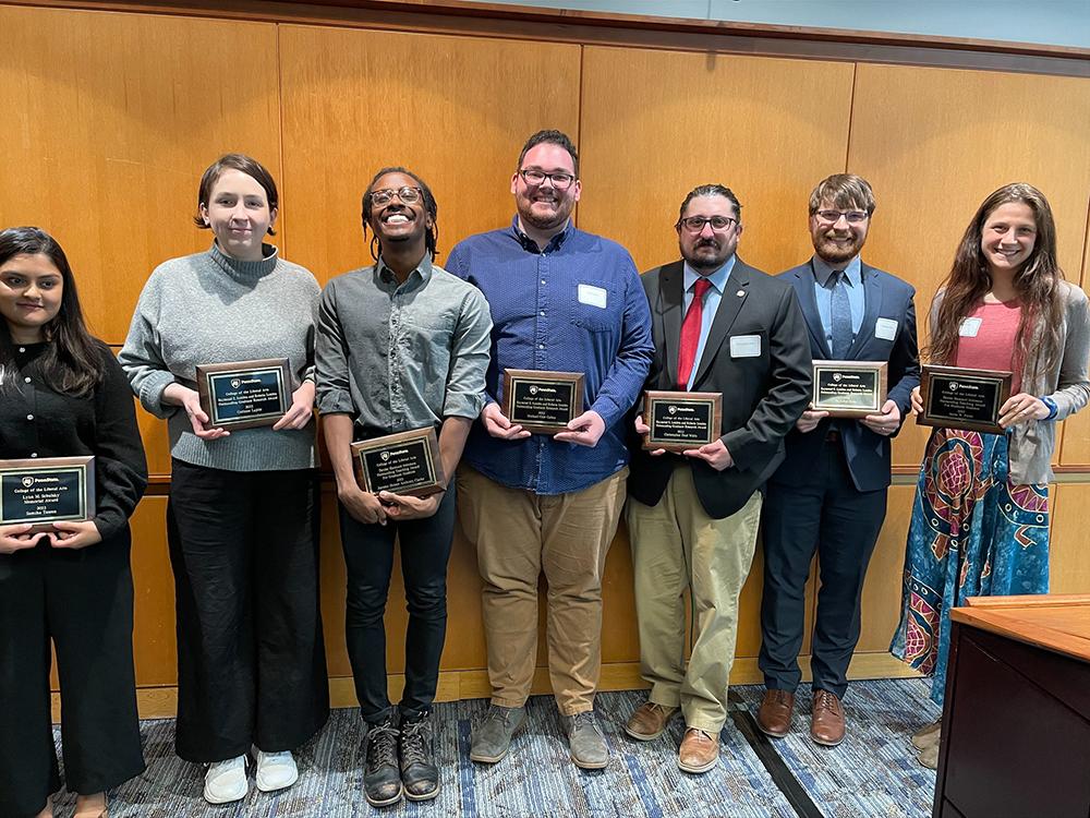 Seven graduate students stand and smile at the camera while each holding an award