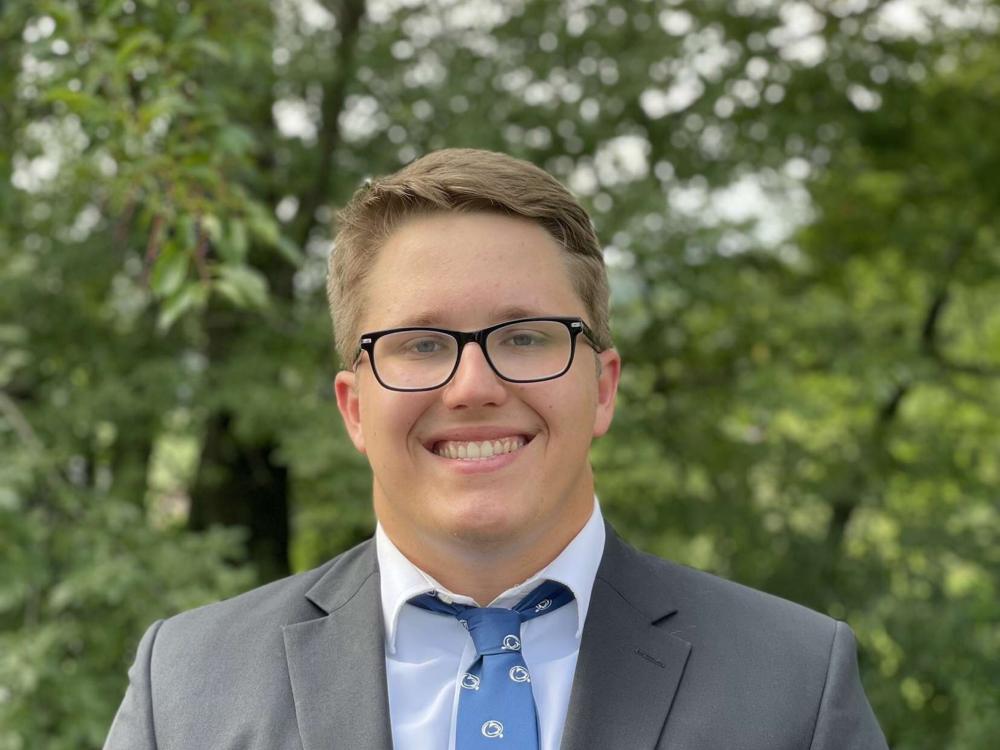 A male student smiling and wearing a suit and tie with trees in the background