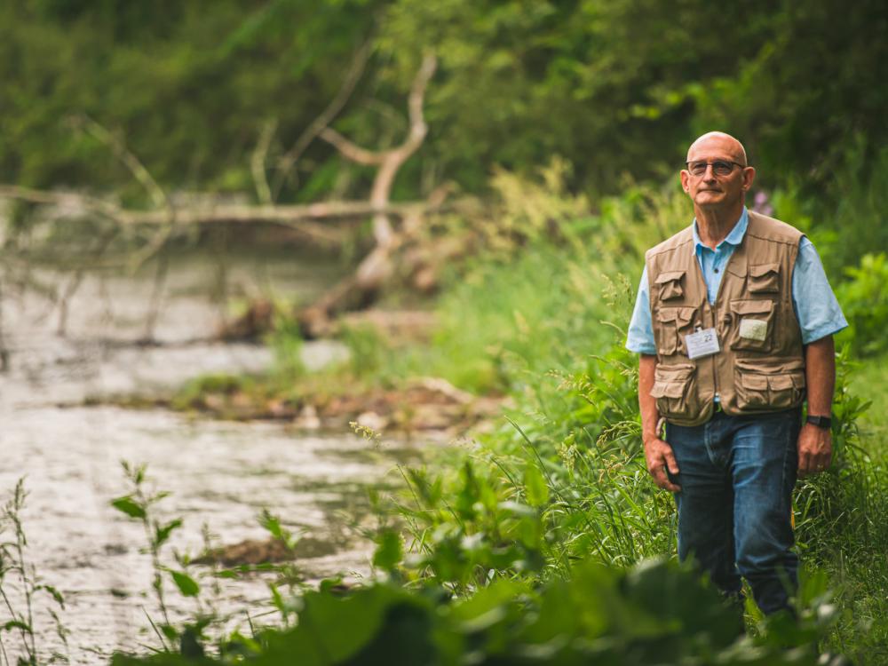 Richard Roush, dean of the College of Agricultural Sciences, stands in fishing gear among greenery beside a flowing creek