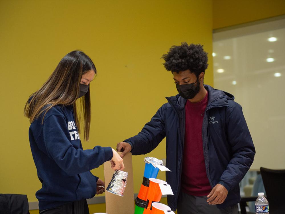 Two students stand in front of a yellow wall and arrange plastic cups and other materials.