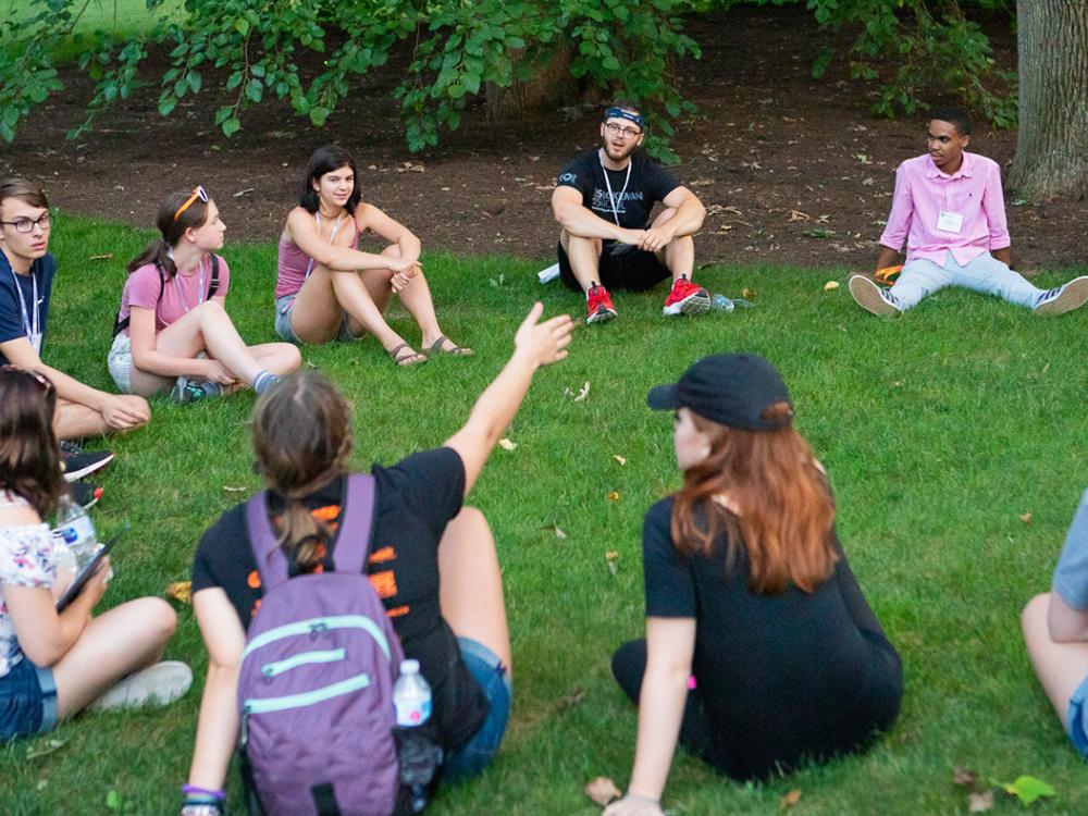 Summer camp students sitting in a circle on a patch of grass and talking.