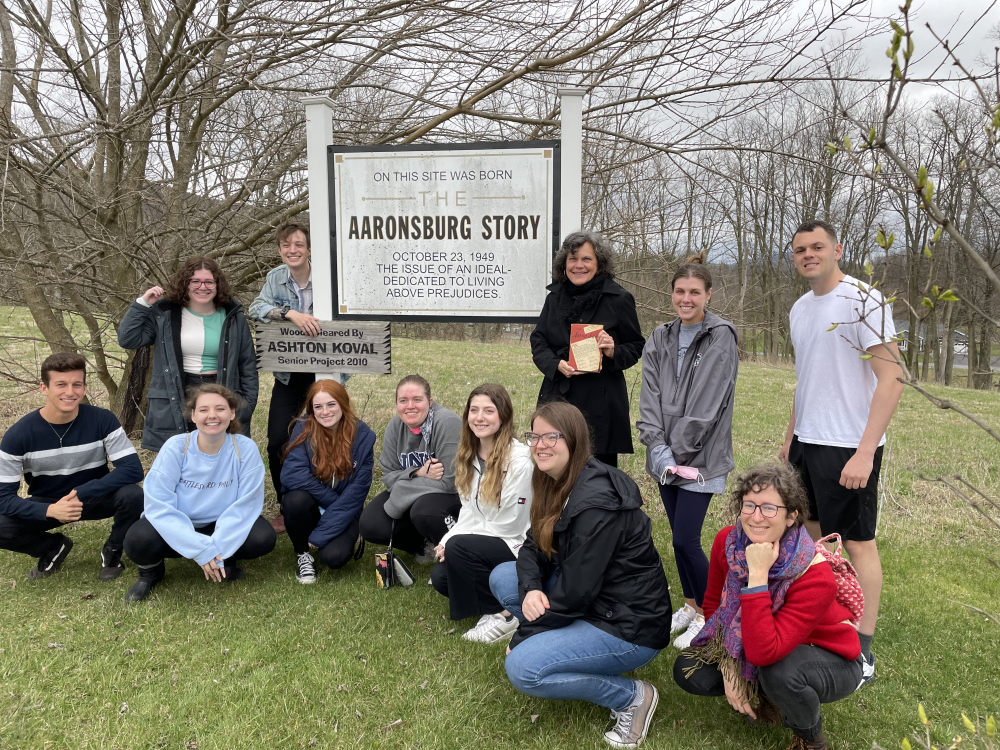 A group of students pose in front of a sign that says "On this site was born The Aaronsburg Story October 23, 1949 The issue of an ideal-dedicated to living above prejudices"