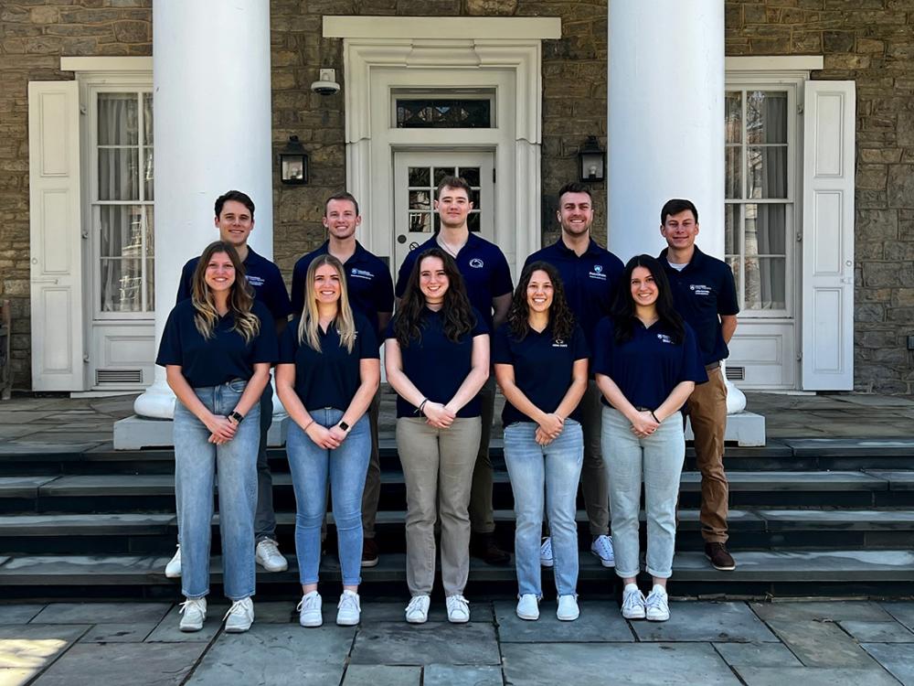 Two rows of five students wear matching blue shirts and stand on steps in front of a beige building. 