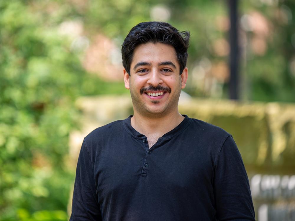 A man smiles at the camera. Greenery is in the background. 