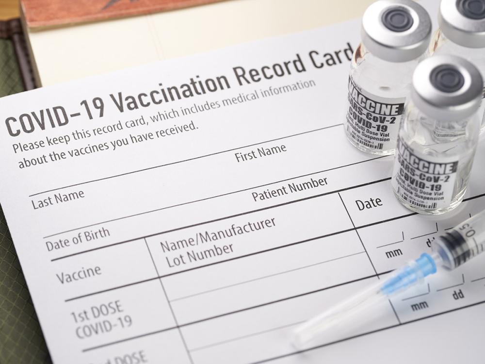 A syringe and COVID-19 vaccine vials sit atop a blank COVID-19 vaccination record card. 