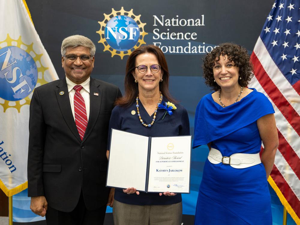 Three people standing in front of a wall with the NSF logo. Kathryn Jablokow is in the middle, holding an award.