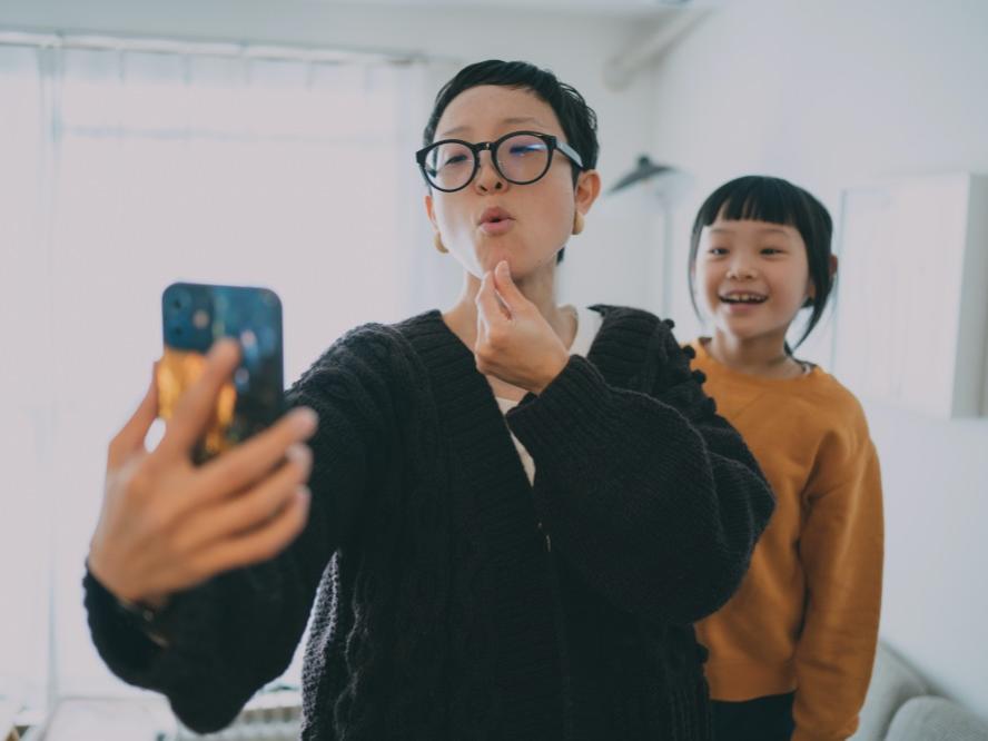 A woman uses sign language to communicate into a smartphone while a child behind her watches and smiles.