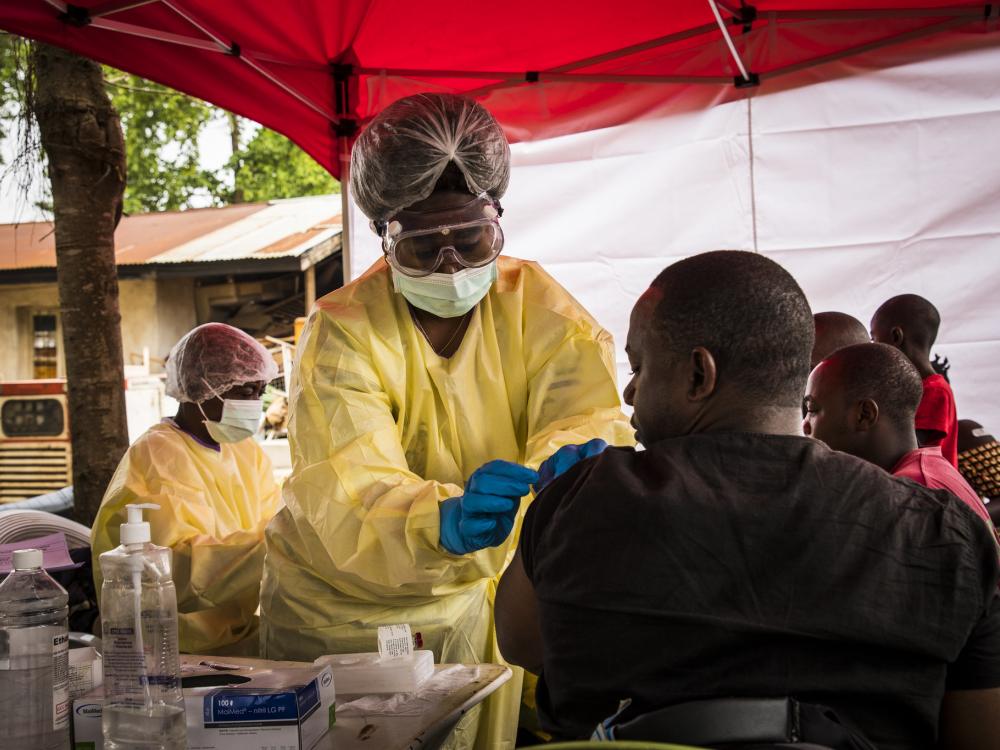 standing woman in protective gear gives vaccine to sitting man