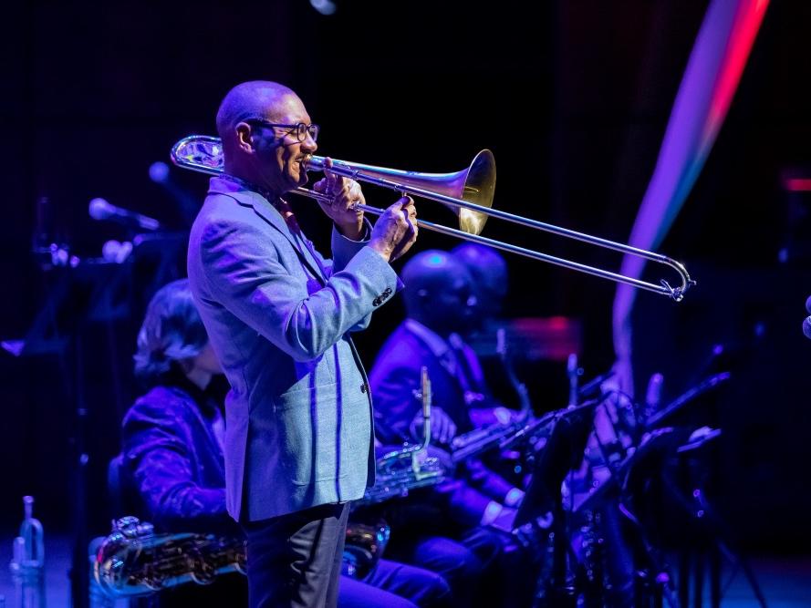 A man wearing eyeglasses and a suit stands and plays a trombone.