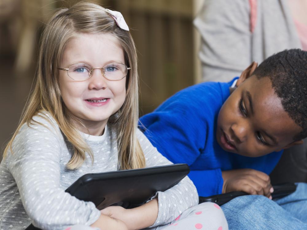 Two preschool students sit together. A White girl holds an iPad while a Black boy looks at her. 