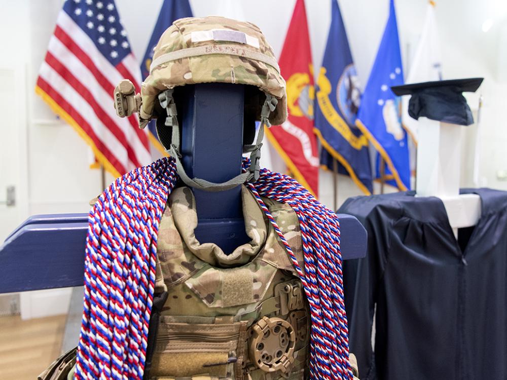 Camouflaged military vest and helmet with red, white and blue honor cords draped over next to blue undergraduate graduation cap and gown.
